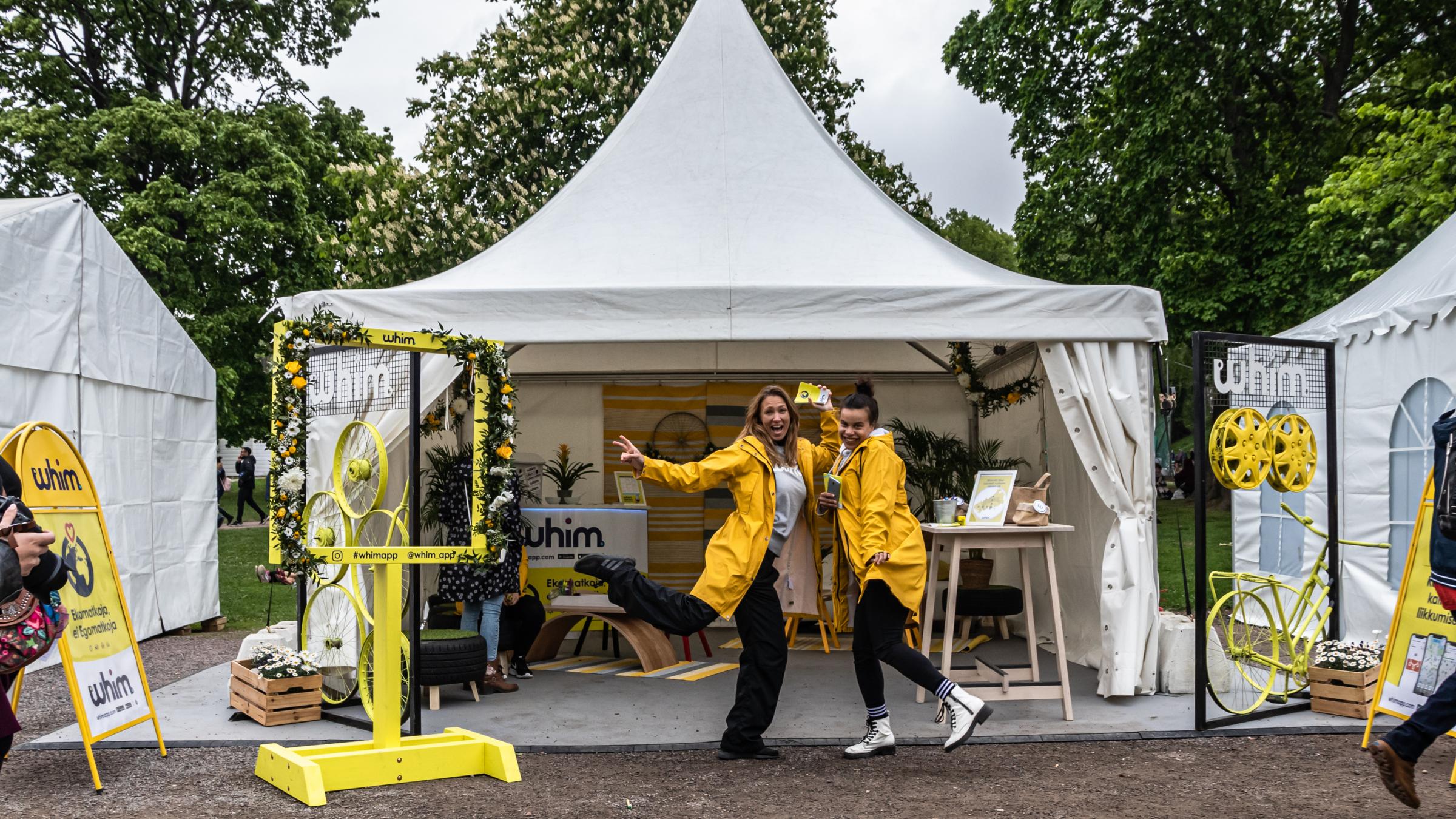 Kuvassa 2 ihmistä keltaisissa sadetakeissa Whim-appin näytteilleasettajapisteellä Maailma kylässä -festivaalilla 2019