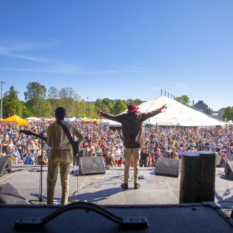 A picture taken from behind Jupiter & Okwess on stage, the artist spreading his hands and a crowd in front of the stage.