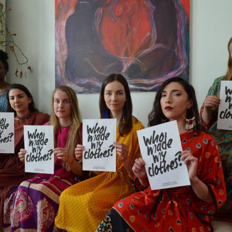 Sohvalla istuvia naisia värikkäissä vaatteissa pitelemässä kylttejä, joissa lukee "Who made my clothes?"