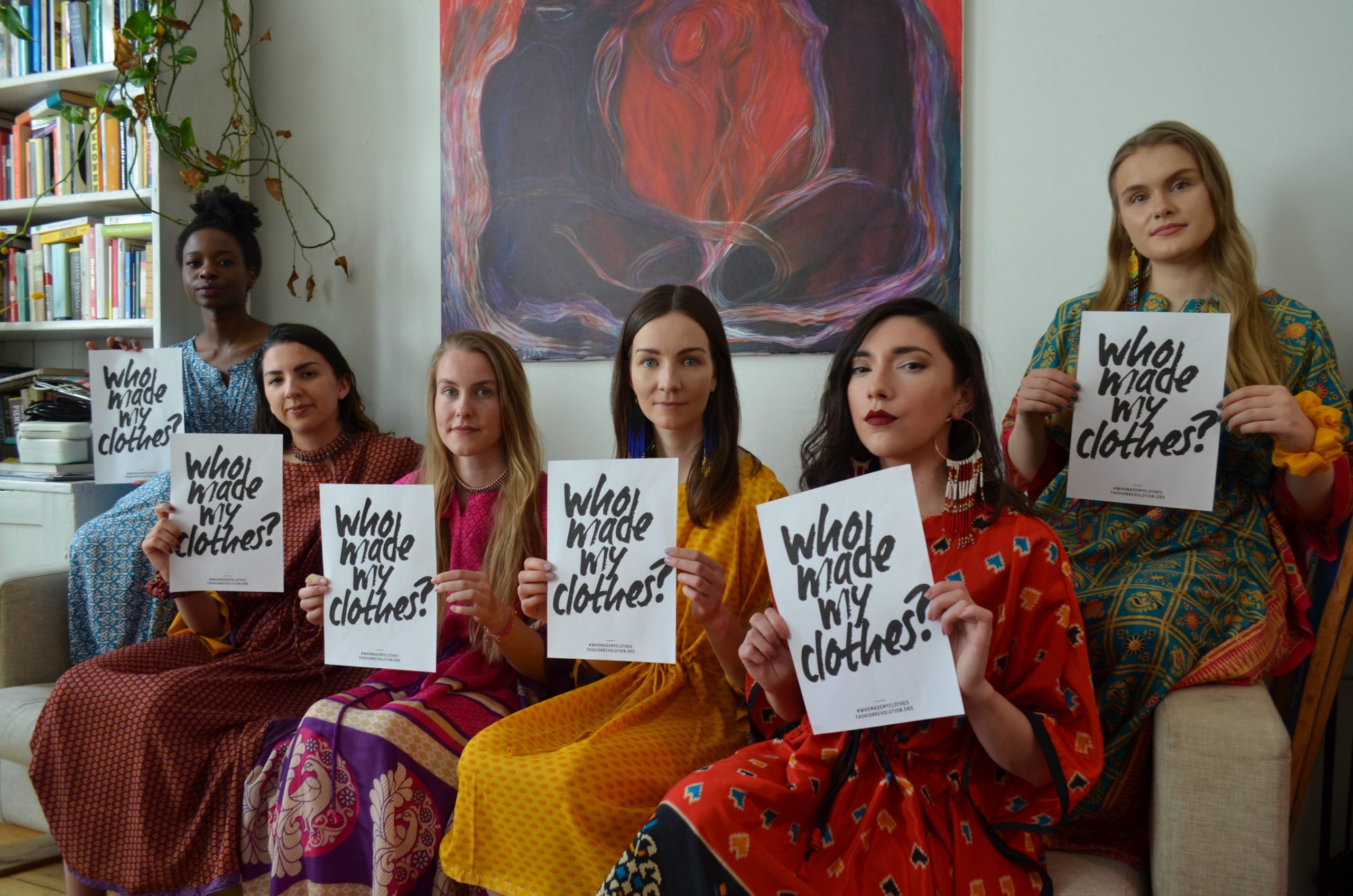 Sohvalla istuvia naisia värikkäissä vaatteissa pitelemässä kylttejä, joissa lukee "Who made my clothes?"