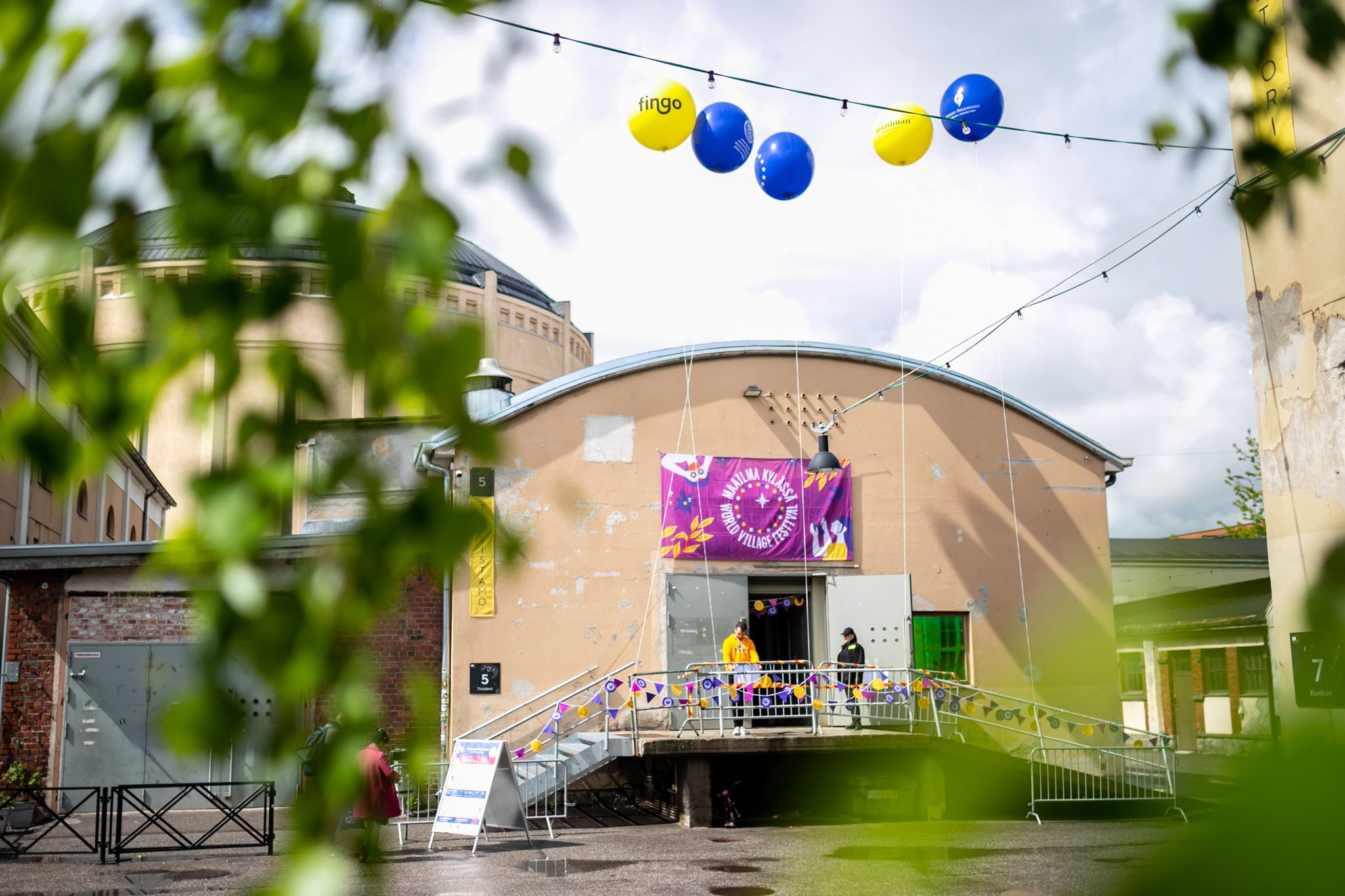 Tiivistämön sisäänkäynti ja Maailma kylässä -festivaalin banneri, sekä sinisiä ja keltaisia ilmapalloja.