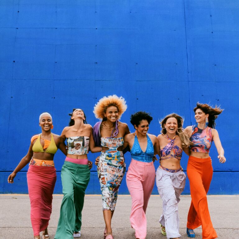 Six women in colorful clothes walk hand in hand, smiling.