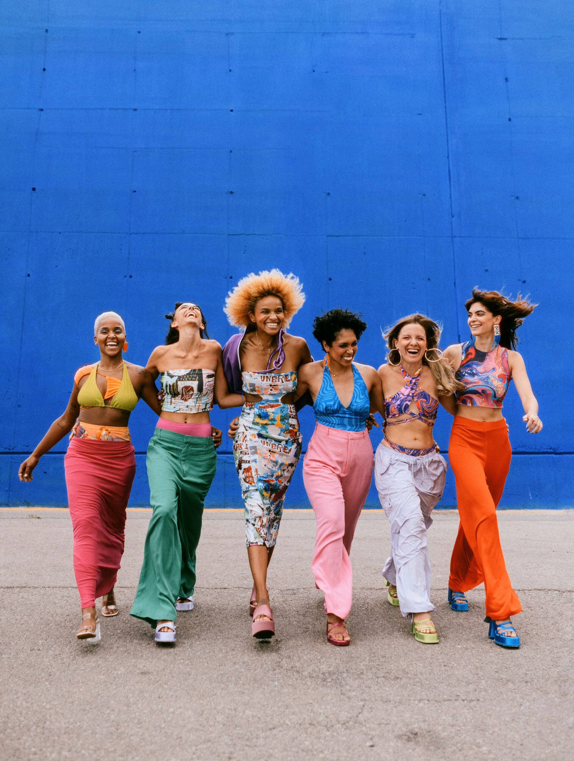 Kuusi naista värikkäissä vaatteissa kävelevät käsi kädessä hymyillen.