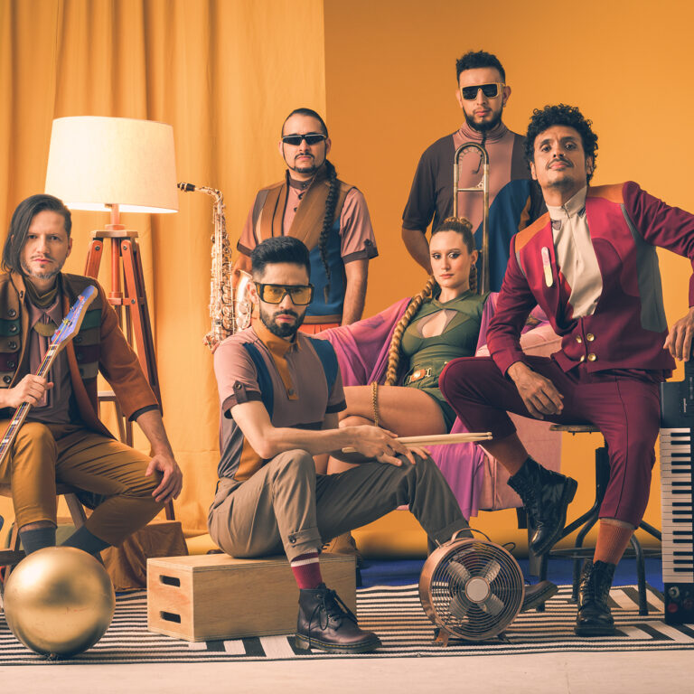 A band of six people with instruments in front of a yellow background.