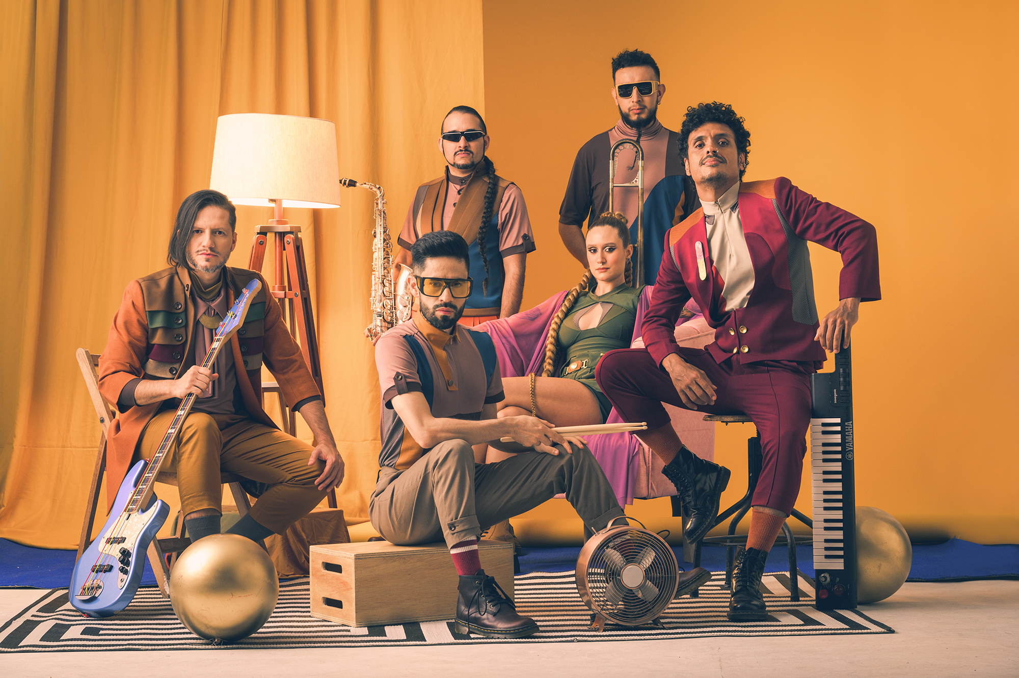 A band of six people with instruments in front of a yellow background.